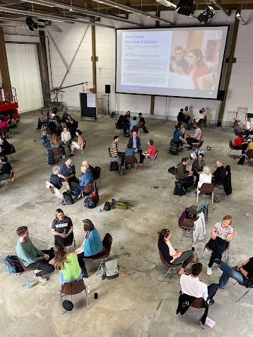 Menschen sitzen in kleinen Gruppen zusammen in Stuhlkreisen und unterhalten sich. 