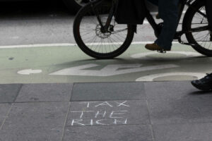 Mit Kreide auf Gehweg geschriebene Worte „TAX THE RICH“, im Hintergrund ein Fahrradfahrer.