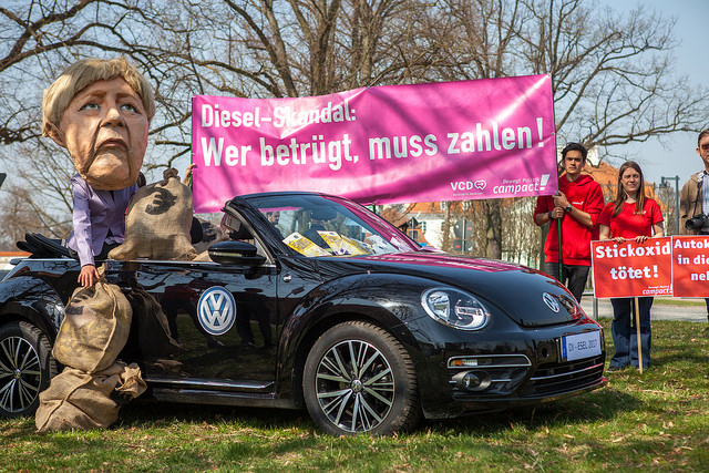 Merkel sitzt in einem VW, dahinter Plakat mit Aufschrift. Diesel-Skandal: Wer betrügt, muss zahlen