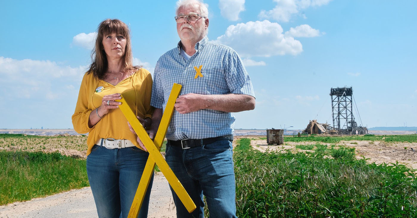 Marita Dresen und Helmut Kehrmann wohnen am Tagebau Garzweiler II in Nordrhein-Westfalen. Sie halten ein großes gelbes X in der Hand. Mit ihrer WeAct-Petition fordern sie: Kein weiteres Dorf mehr für Kohle: Unterzeichnen Sie jetzt für Klimagerechtigkeit hier und überall