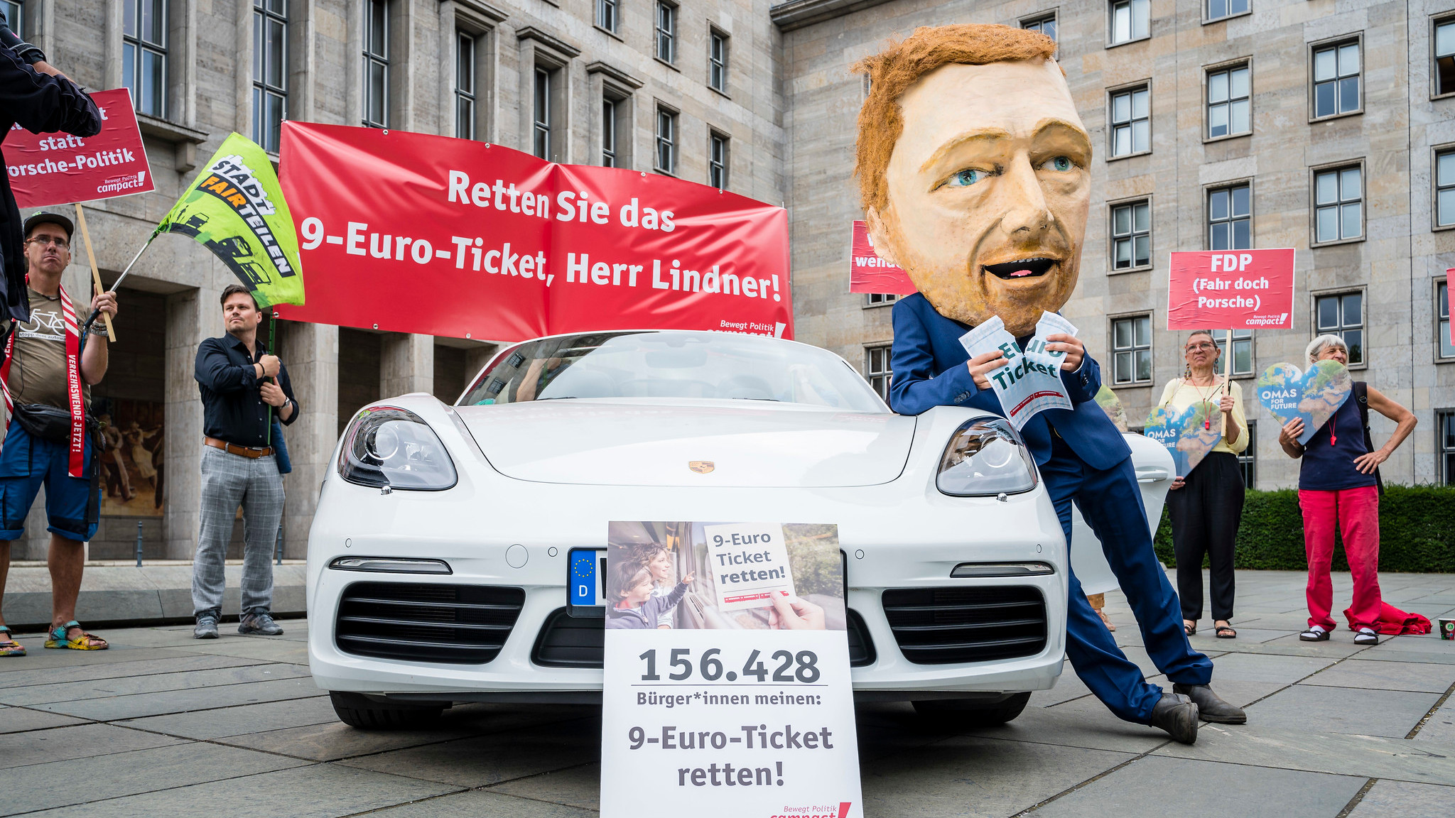 Eine Person trägt eine übergroße Nachbildung von Bundesfinanzminister Christian Lindner's Kopf und lehnt sich, beim zerreißen eines 9-Euro-Tickets, an ein Porsche-Cabrio. Vor dem Auto steht der Appell "9-Euro-Ticket retten!".