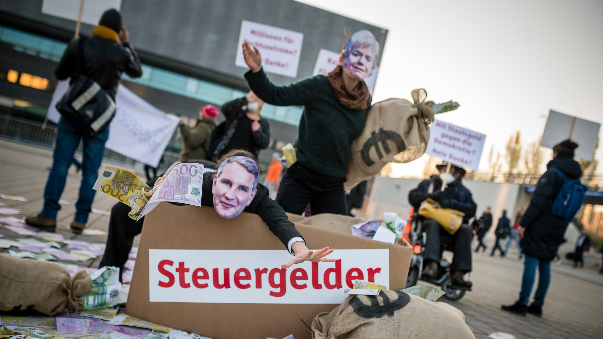 Zwei Personen mit Masken von Erika Steinbach und Björn Höcke werfen Geldscheine. Daneben sind Säcke mit einem Euro-Zeichen, sowie ein Karton auf dem "Steuergelder" steht.