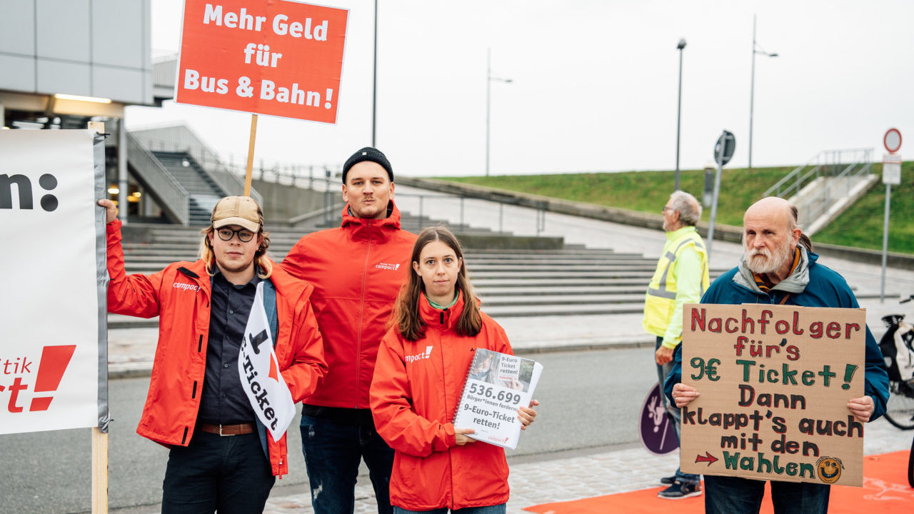 Campact-Aktivist*innen demonstrieren am Rande der Verkehrsministerkonferenz. Sie halten Schilder und die gebundenen Unterschriftensammlung zum Campact-Appell "9-Euro-Ticket retten!", sowie ein Schild aus Pappe mit der Aufschrift: "Nachfolger fürs 9€ Ticket! Dann klappt's auch mit den Wahlen"