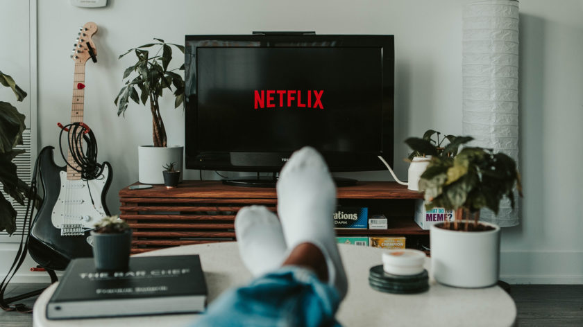 Eine Person sitzt auf einem Sofa, das Foto ist aus ihrer Perspektive aufgenommen, ihre Füße liegen auf einem Tisch. Sie schaut auf einen Fernseher, auf dem das Netflix-Logo zu sehen ist.