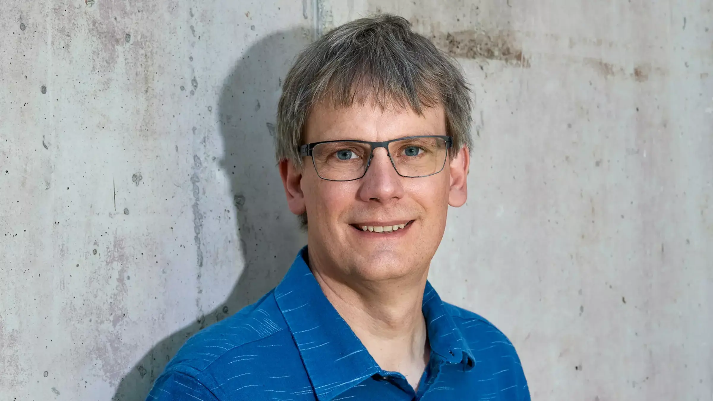 Felix Kolb trägt eine Brille und ein dunkel blaues Hemd. Er lächelt und steht vor einer grauen beton Wand.