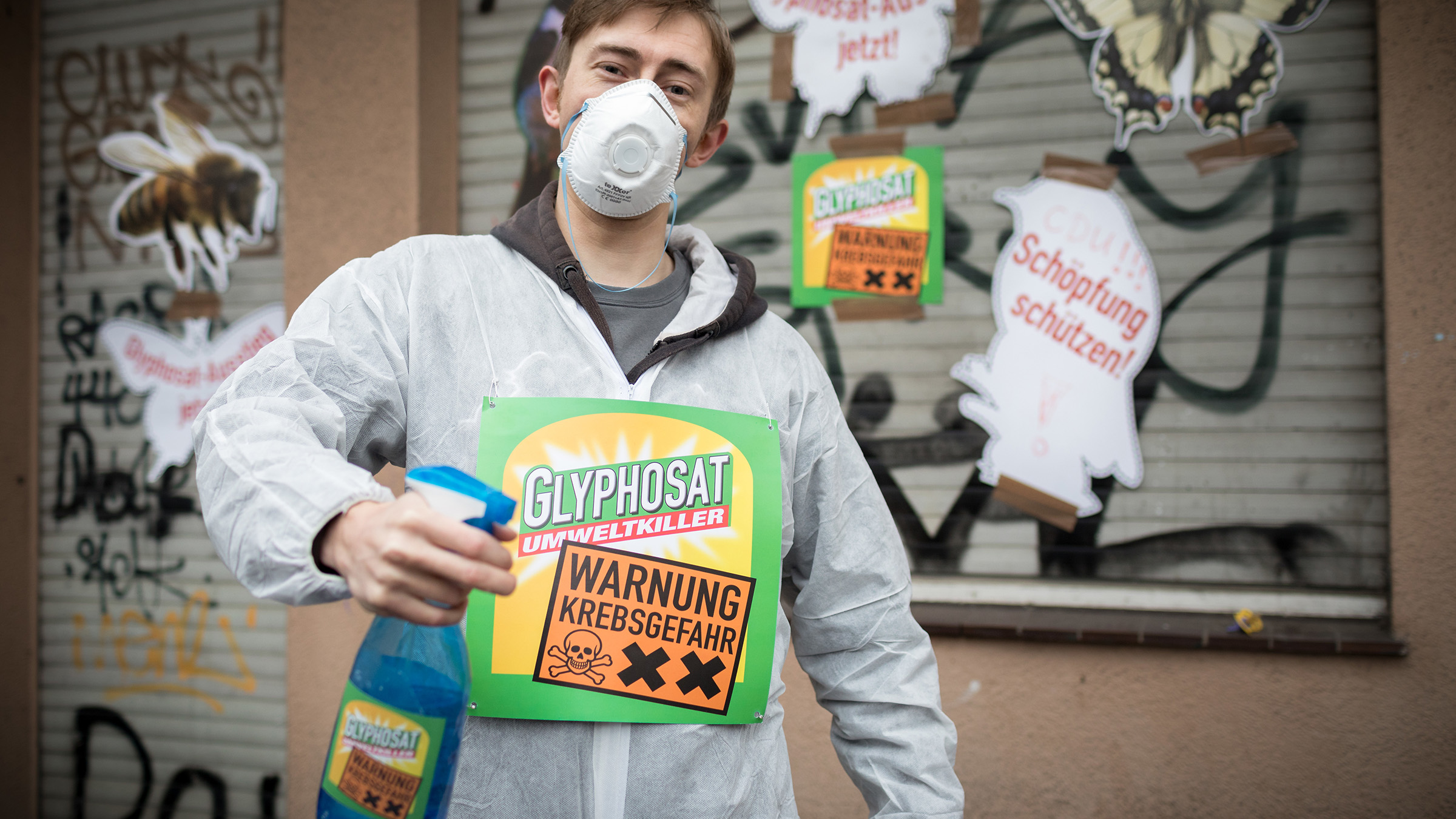 Ein Aktivist im Schutzanzug mit einer Glyphosat-Spritzflasche in der Hand. Darauf die Warnung: Krebsgefahr