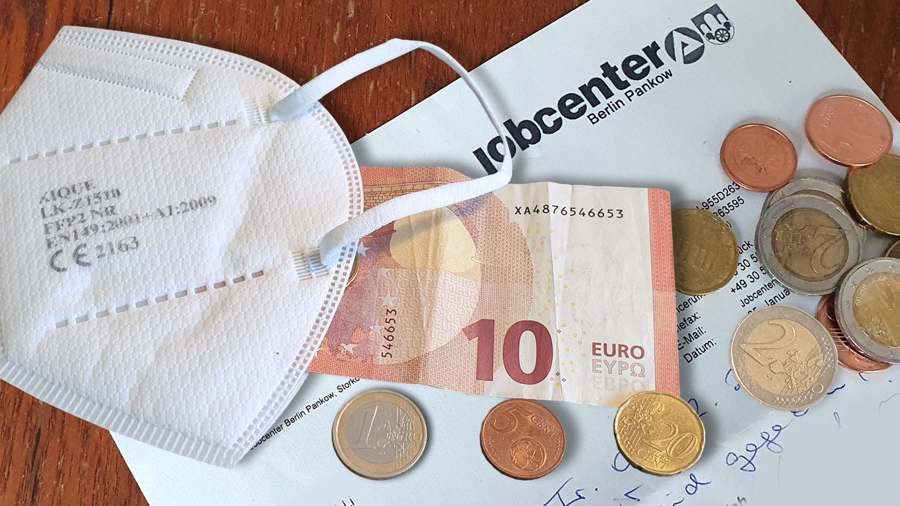 Ein Bescheid vom Jobcenter, darauf liegt ein Zehn-Euro-Schein und einige Münzen sowie eine Maske.