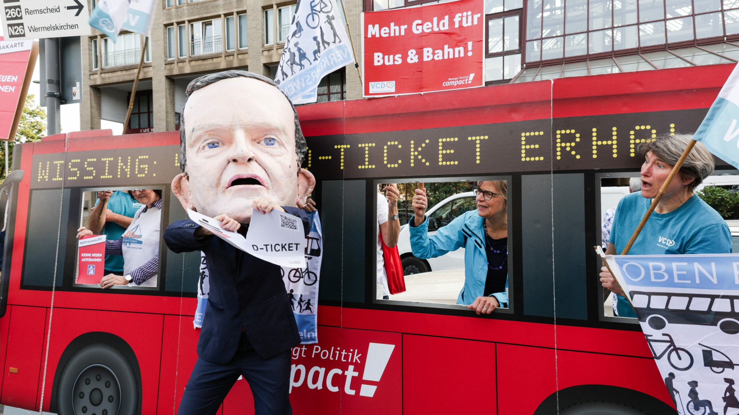 Ein Wissing-Schauspieler zerreißt das Deutschlandticket vor Demonstrierenden in einem Papp-Bus.