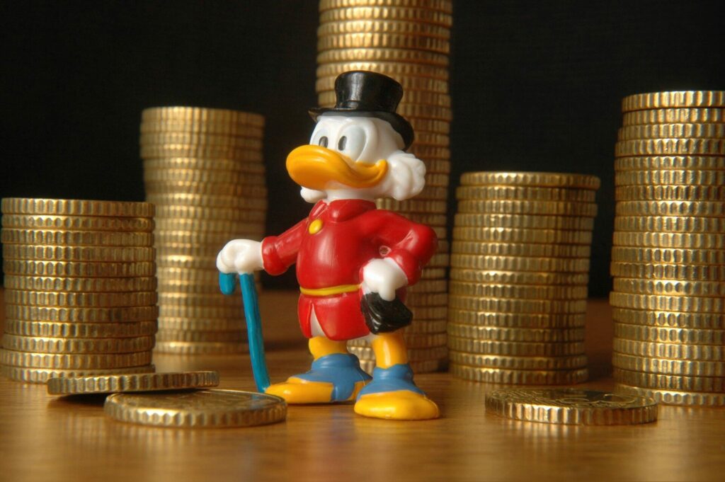 Das Bild zeigt eine Plastikfigur von Dagobert Duck, die vor aufgetürmten goldenen Münzstapeln steht.