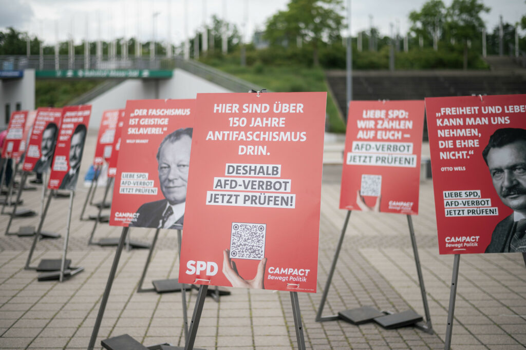 Reihe großformatiger Plakate mit Porträts und Zitaten von SPD-Politikern wie Willy Brandt und Otto Wels. Die Plakate stehen vor dem Eingang des SPD-Parteitags in Berlin. Aufschriften fordern die SPD auf, ein AfD-Verbotsverfahren zu prüfen.