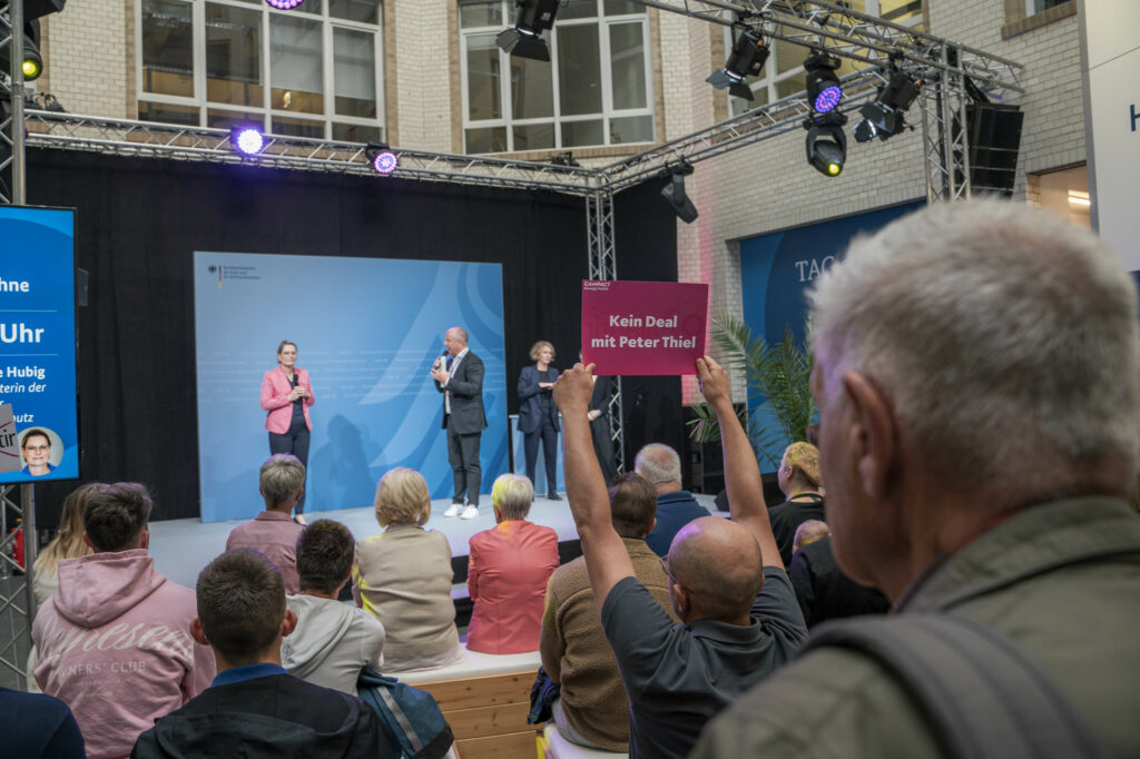 Bei einer Diskussionsveranstaltung hebt ein Teilnehmer ein Schild mit der Aufschrift „Kein Deal mit Peter Thiel“ hoch. Auf der Bühne sprechen eine Frau im rosa Blazer und ein Mann im Anzug vor dem Publikum.