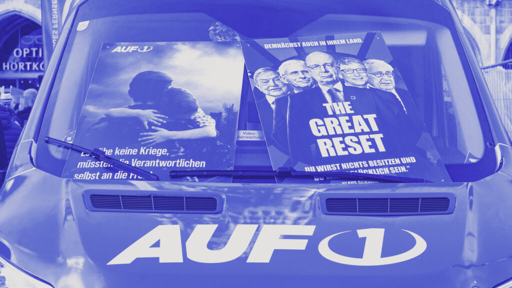 Blauer AUF1-Transporter mit zwei großformatigen AUF1-Plakaten auf der Windschutzscheibe; das linke Poster zeigt eine Umarmung als emotionales Motiv, das rechte thematisiert den „Great Reset“ als typisches Narrativ des Verschwörungssenders.