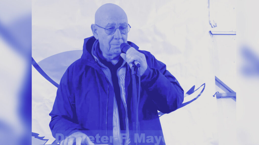 Dr. Peter F. Mayer spricht auf der Kundgebung „Neutralität schafft Frieden!“ am 6. Januar 2024 am Platz der Menschenrechte in Wien in ein Mikrofon; er trägt eine dunkle Jacke mit grüner Innenseite und steht hinter einem runden Rednerpult mit Manuskript, vor einem weißen Banner im Hintergrund.