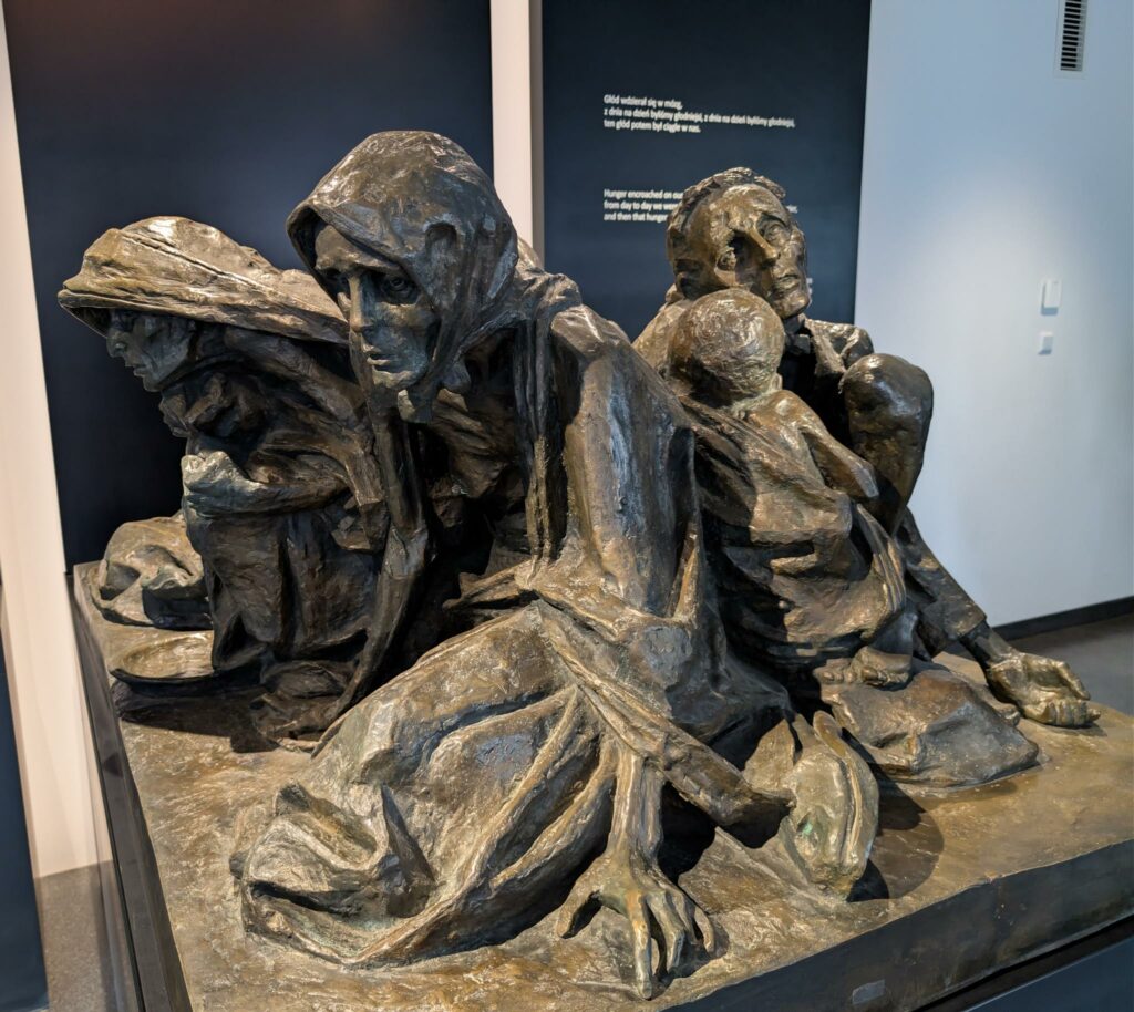 Eine Skulptur in der Gedenkstätte Auschwitz-Birkenau zeigt eine Gruppe ausgemergelter und kraftloser Frauen und Kinder, die am Boden kauernd den Tod erwarten.