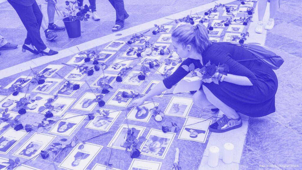 Auf dem Bild sieht man einige Fotos der 77 Opfer des rechtsextremistischen Anschlags in Norwegen 2011, die zum Gedenken auf dem Gehweg ausgelegt wurden. Eine Frau legt eine Blume nieder, hinter ihr stehen etwas verlorene und bedrückt weitere Trauernde.