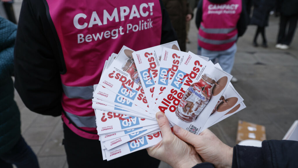 Nahaufnahme einer Hand, die einen Fächer aus Campact-Stickern hält. Auf den Stickern steht „Alles AfD oder was?“, daneben ist eine Müllermilch-Flasche abgebildet. Im Hintergrund stehen Aktive in pinken Campact-Westen mit der Aufschrift „Campact – Bewegt Politik“.